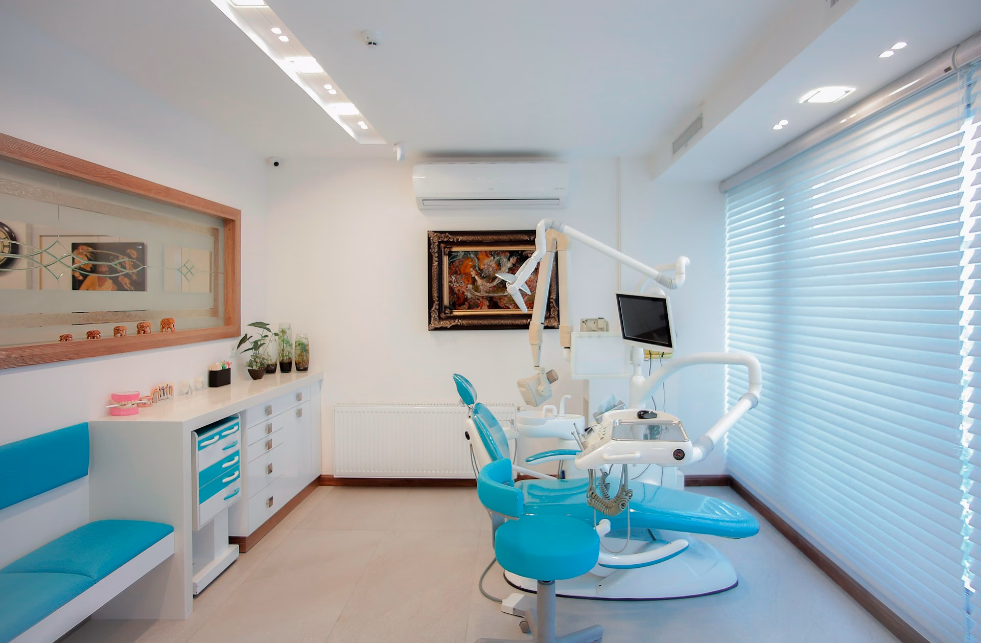 A modern dental office with turquoise chairs, dental equipment, a computer screen, cabinets, wall art, and large windows with blinds letting in natural light. The space is clean, bright, and organized.