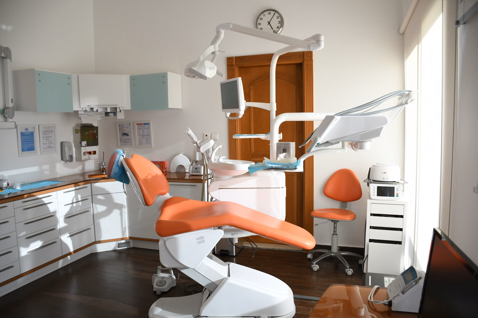A modern dental clinic with an orange dental chair, dental equipment, cabinets, and a clock on the wall. Sunlight streams in through a window, brightening the clean, organized room.