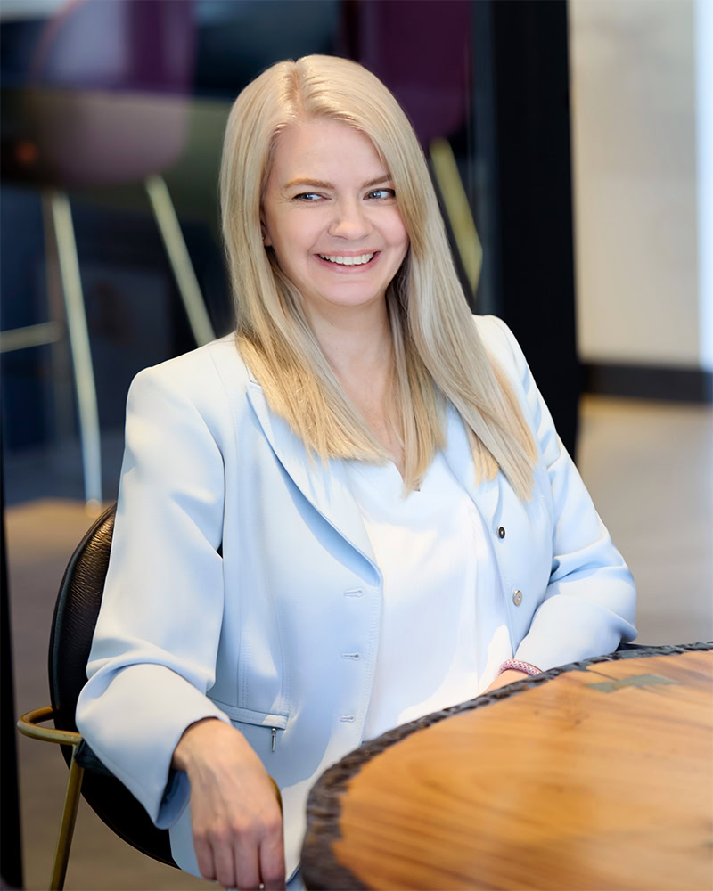 A woman with long blonde hair, wearing a light blue blazer and white blouse, sits at a wooden table and smiles at the camera in a modern office setting.