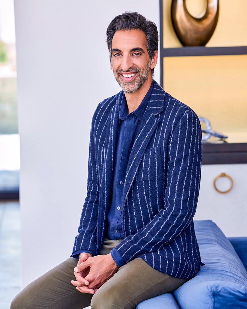 A man with dark hair and a beard, wearing a blue shirt and a blue striped blazer, sits on a blue cushion indoors, smiling at the camera. Shelves with decorative items appear in the background.