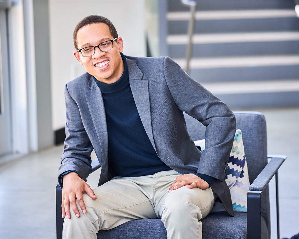 A man wearing glasses, a gray blazer, and light pants sits smiling on a modern chair indoors, with a patterned pillow beside him and stairs visible in the background.
