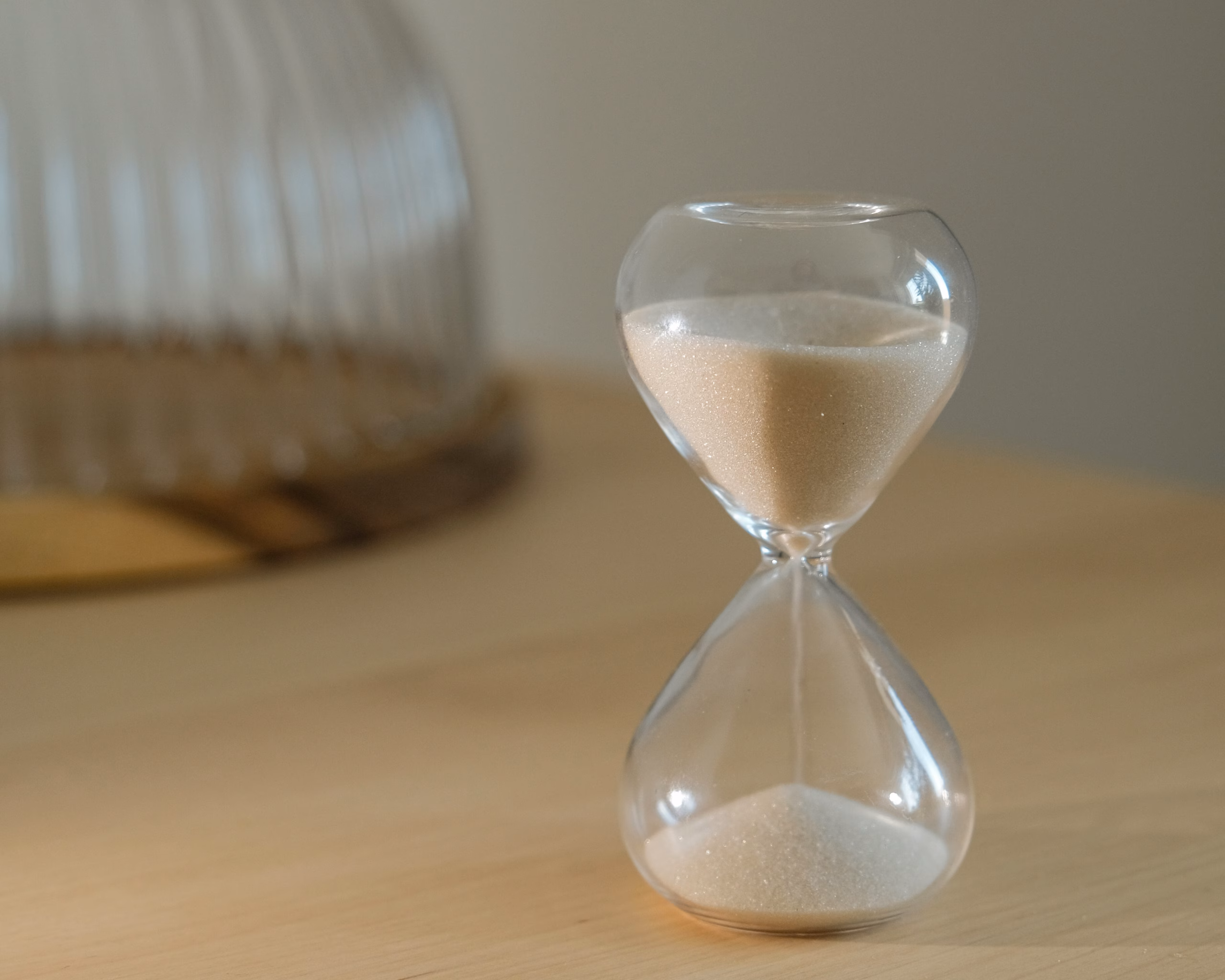 A clear glass hourglass filled with white sand sits on a light wooden surface, with some sand collected at the bottom and some in the top chamber. A blurred object is visible in the background.