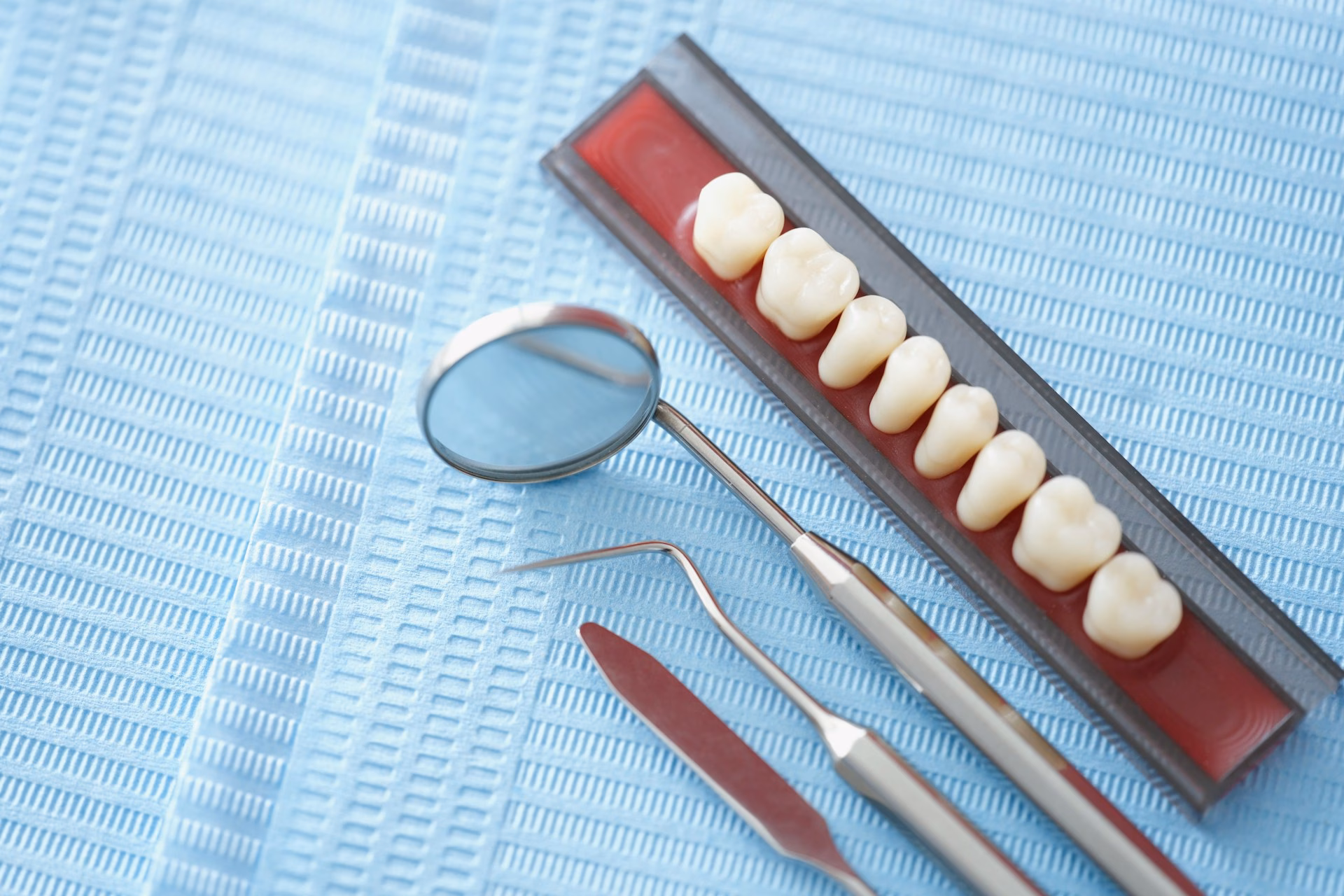 A dental mirror, explorer, scaler, and a row of artificial teeth are arranged on a blue disposable dental bib.